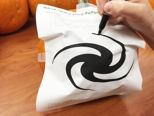 A black marker traces the outline of the template for a galaxy on the pumpkin