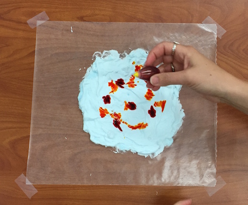 a photo of shaving cream on wax paper and a hand dropping red and yellow food coloring onto it